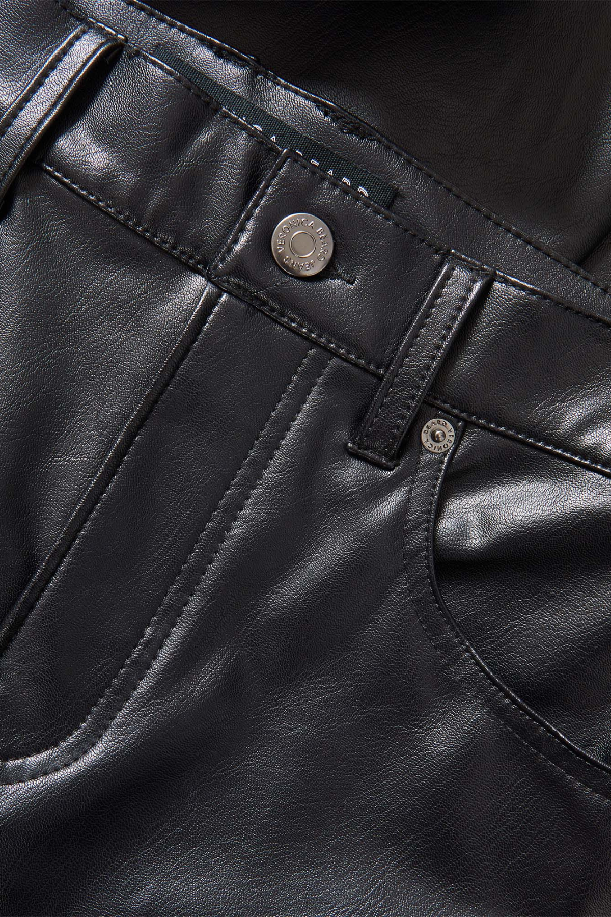 Veronica Beard Beverly High Rise Skinny Flare Pant in black vegan leather, featuring a high-rise waist, front patch pockets and a skinny-flare silhouette. Close-up of black leather pants showing the waistband, belt loops, button closure, and front pocket stitching.